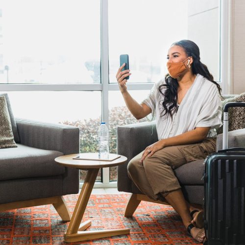 A happy female business traveler video chats as she waits for ride share to the airport. She is wearing a protective face mask as she travels during COVID-19.