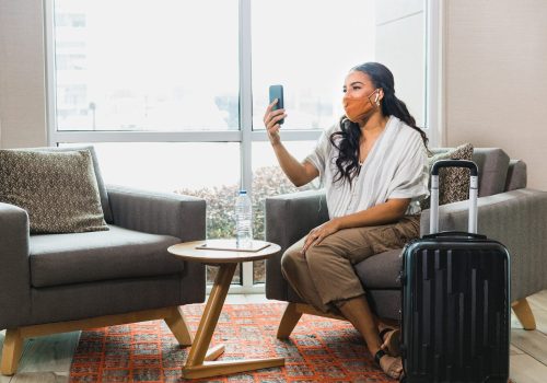 A happy female business traveler video chats as she waits for ride share to the airport. She is wearing a protective face mask as she travels during COVID-19.